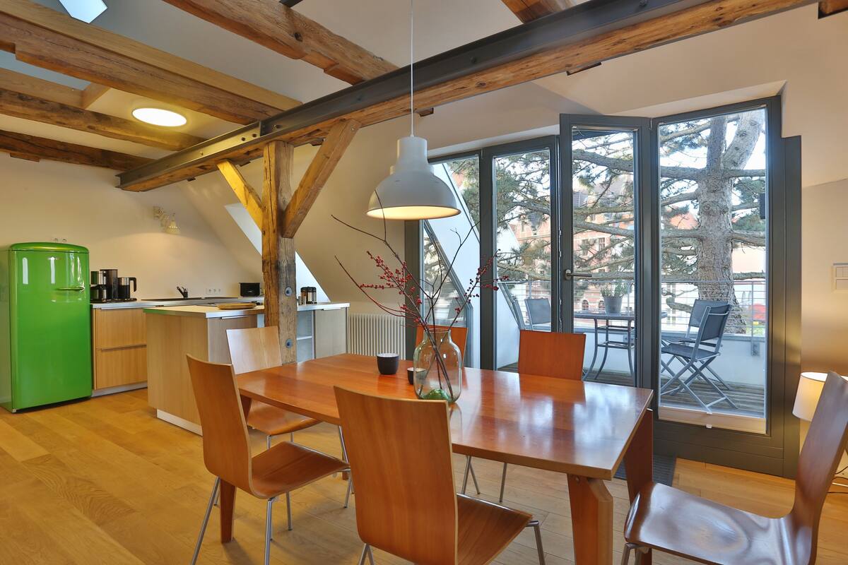 Una cocina moderna con vigas de madera, un comedor con mesa y sillas de madera, y un amplio ventanal con vistas a árboles.
