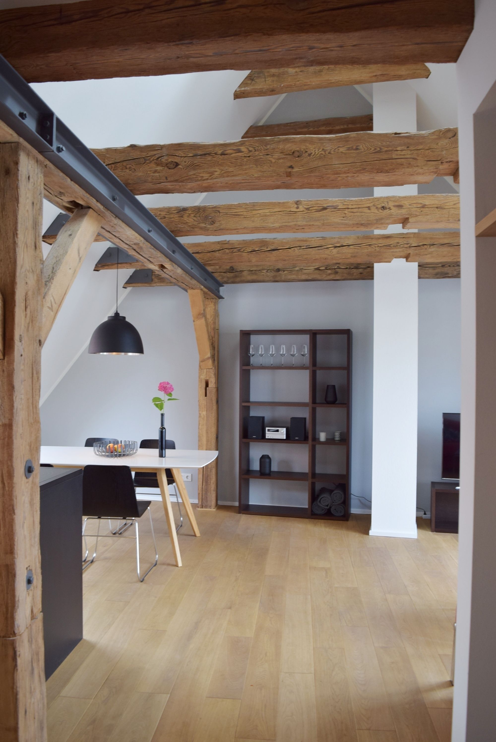Zona pranzo con tavolo bianco, sedie nere e lampadario nero a sospensione. Si nota una libreria in legno con diversi oggetti e un pilastro bianco.