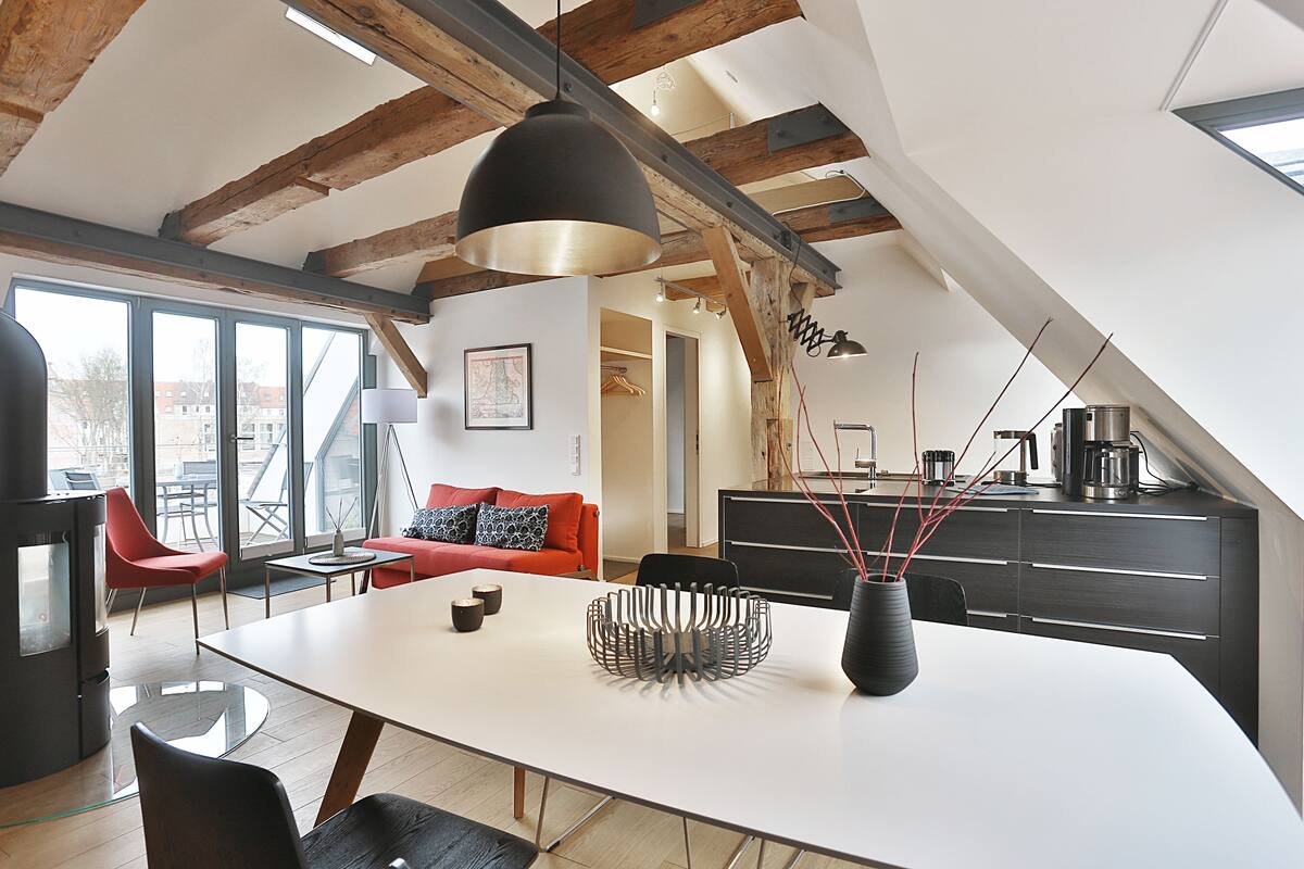 Un loft moderno con vigas de madera a la vista, una zona de comedor con mesa blanca y sillas negras, un sofá rojo y una cocina con armarios oscuros.