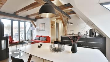 En moderne loftrum med synlige træbjælker, et spiseområde med et hvidt bord og sorte stole, en rød sofa og et køkken med mørke skabe.