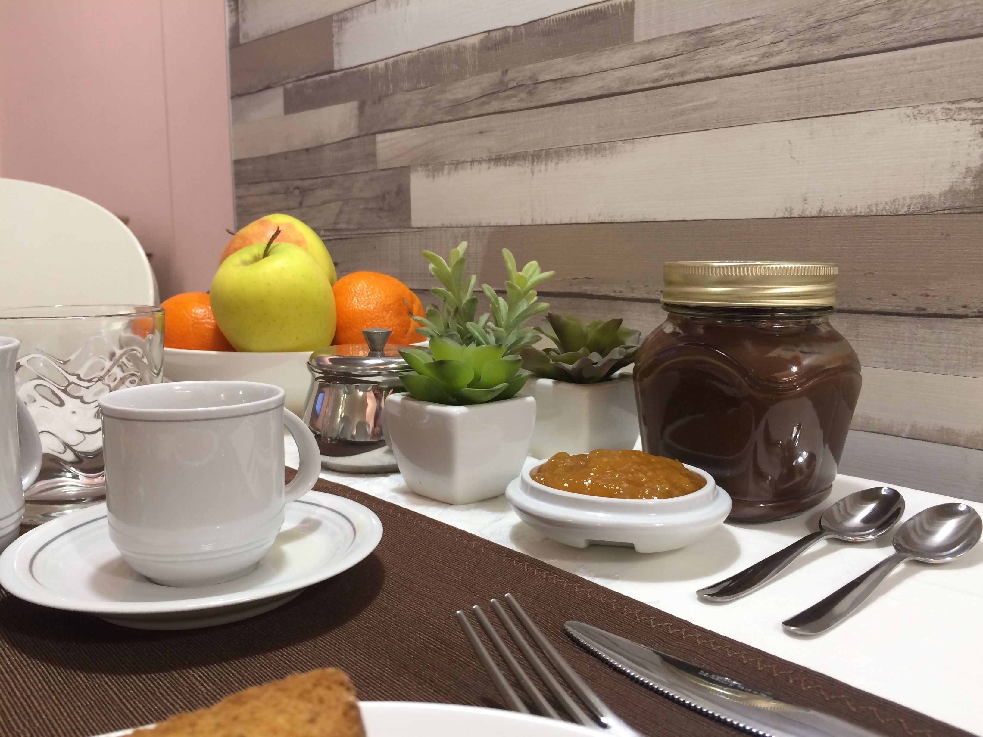 Una mesa de desayuno con tostadas, un vaso de agua, una taza, un frasco de mermelada, un cuenco pequeño de mermelada, una planta en maceta y un bol de fruta.