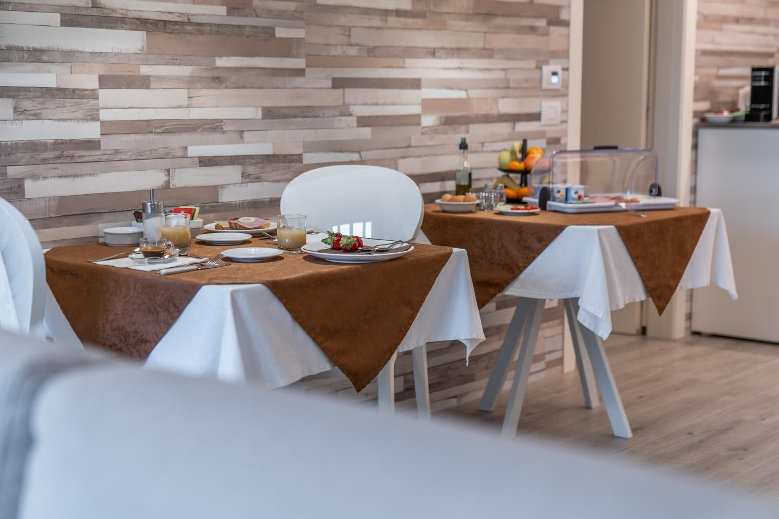 A dining area with two tables set for breakfast, featuring white chairs, a wooden wall, and a kitchen in the background.