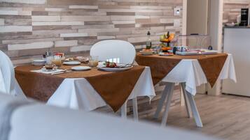 A dining area with two tables set for breakfast, featuring white chairs, a wooden wall, and a kitchen in the background.