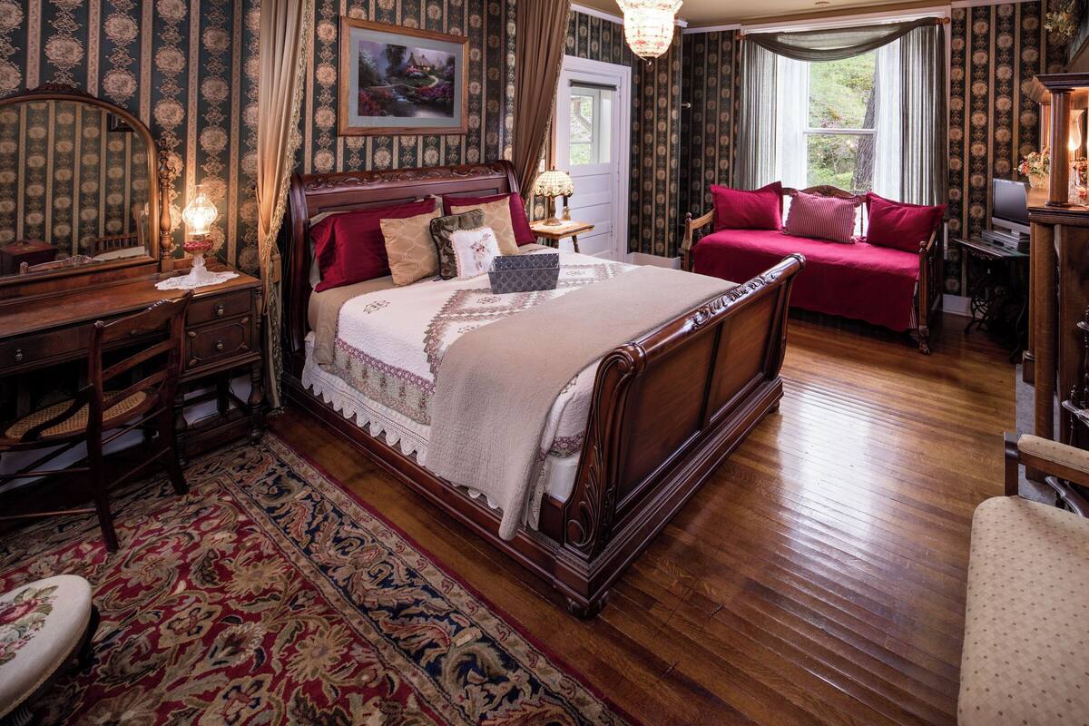 A traditional bedroom with a large bed, a wooden dresser, a red sofa, and a desk with a lamp.