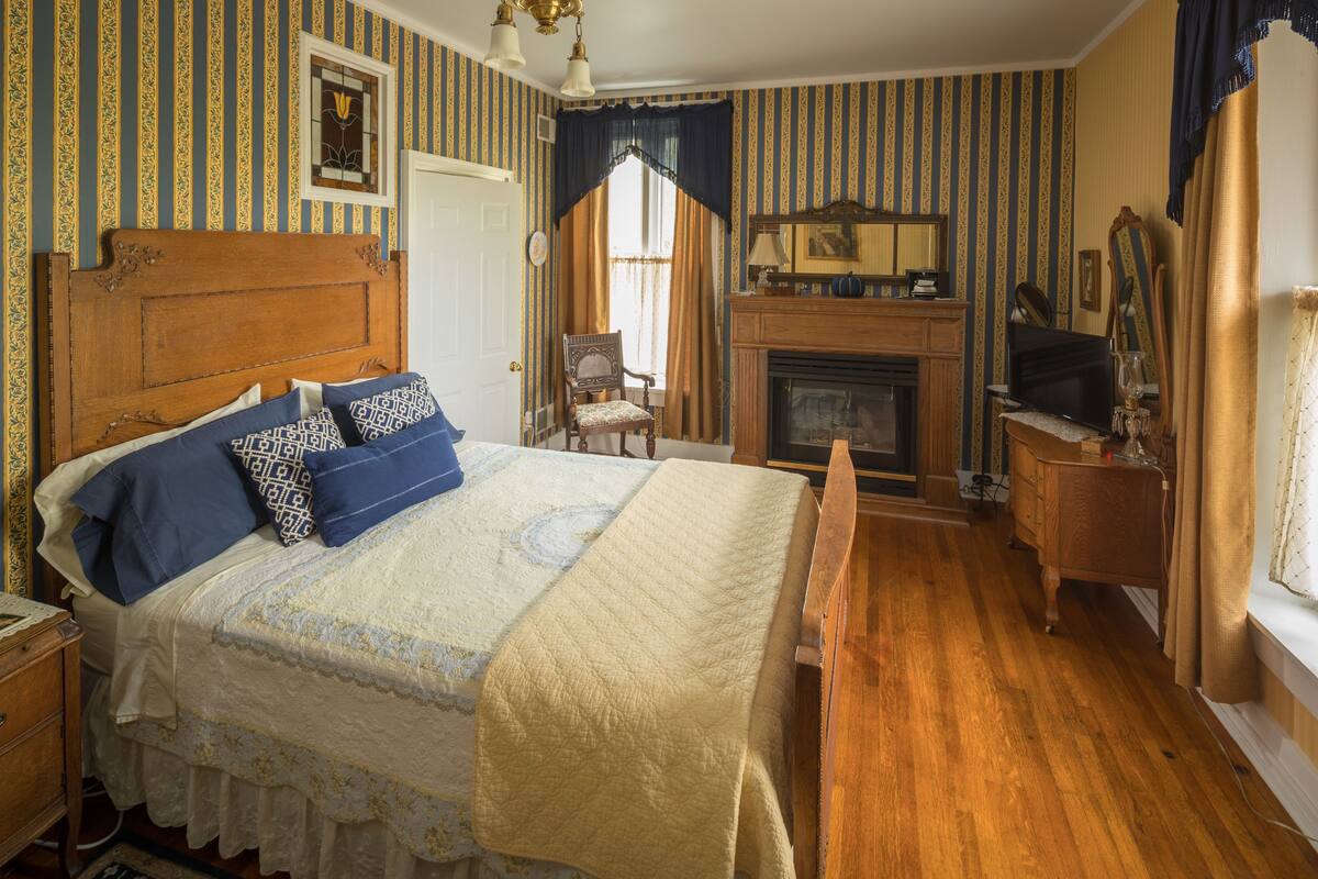 A bedroom with a large bed, a wooden dresser, a chair, a fireplace, and a mirror.