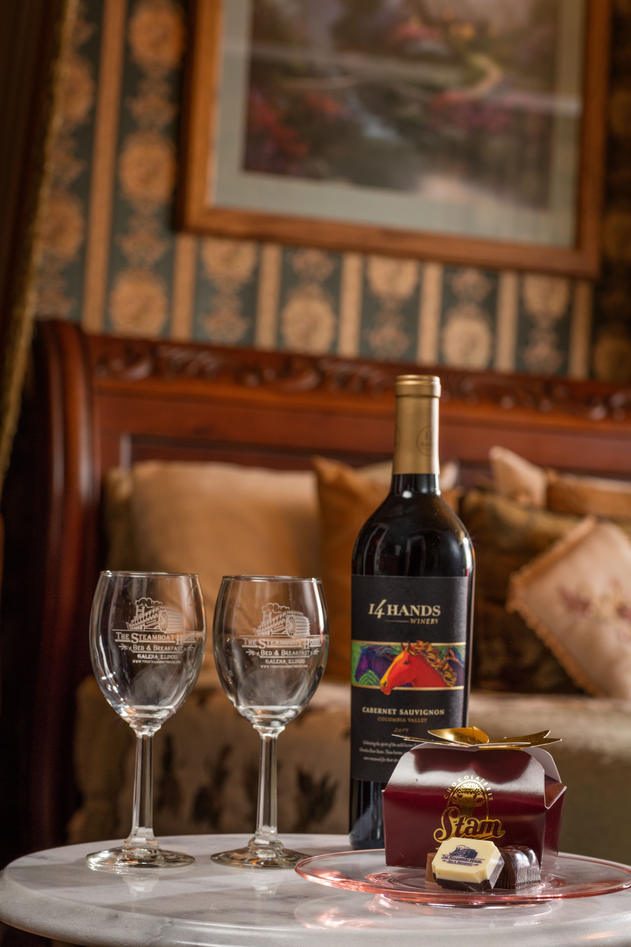 A table with two wine glasses, a bottle of wine, and a dessert on a marble table in a room with patterned wallpaper and a wooden headboard.