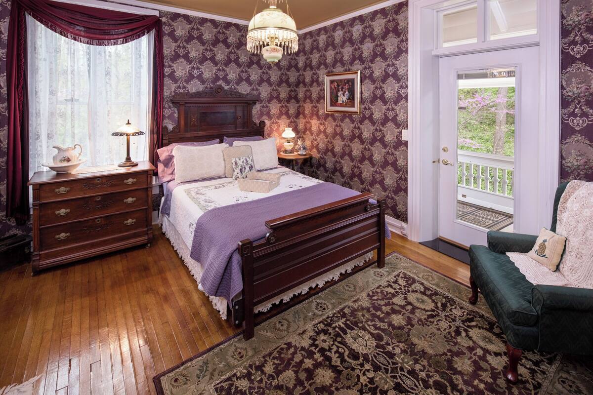 A bedroom with a wooden bed, a dresser, a nightstand, a chandelier, and a view of the outdoors through a door.