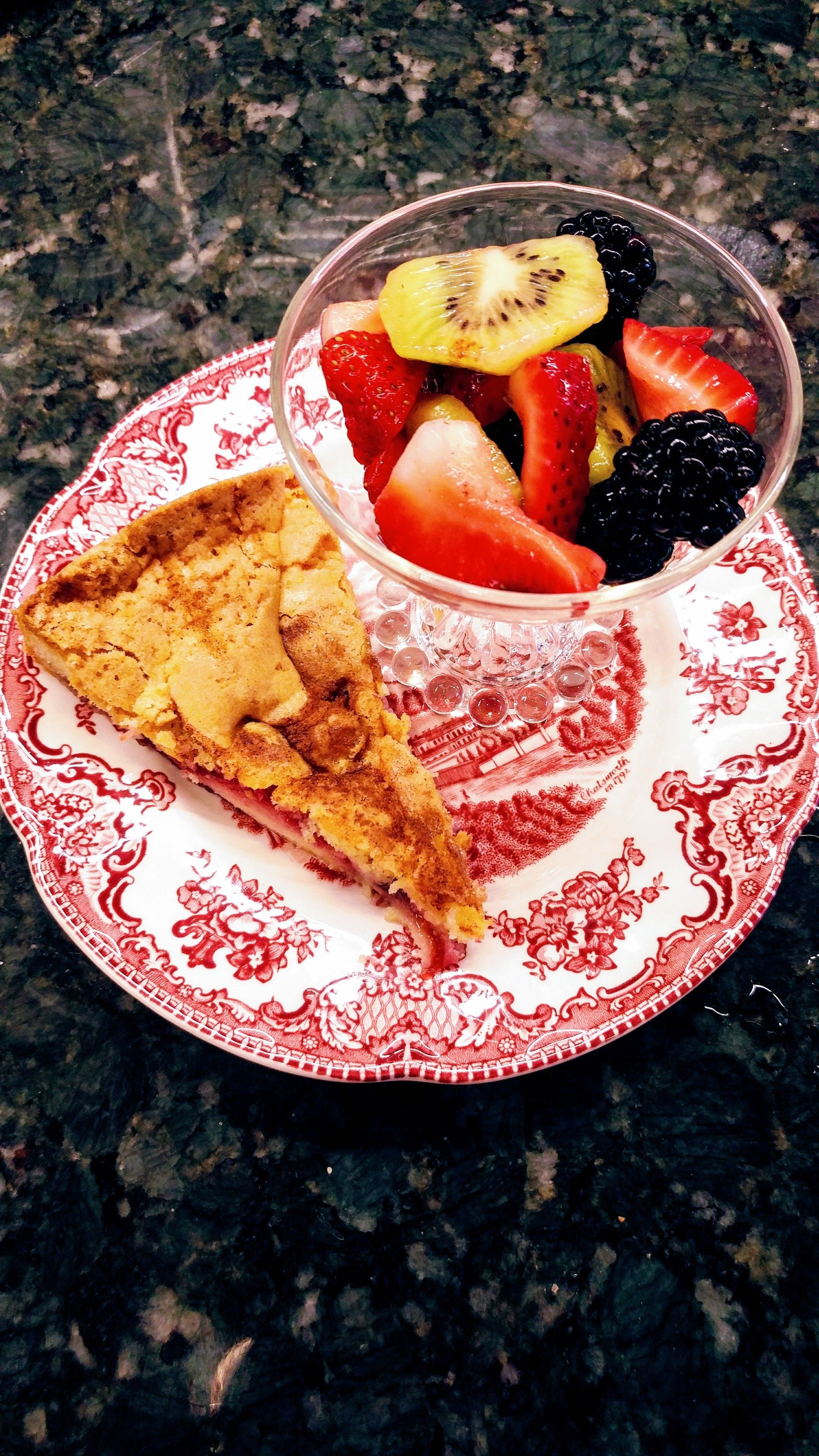A slice of pie on a patterned plate with a fruit bowl beside it.