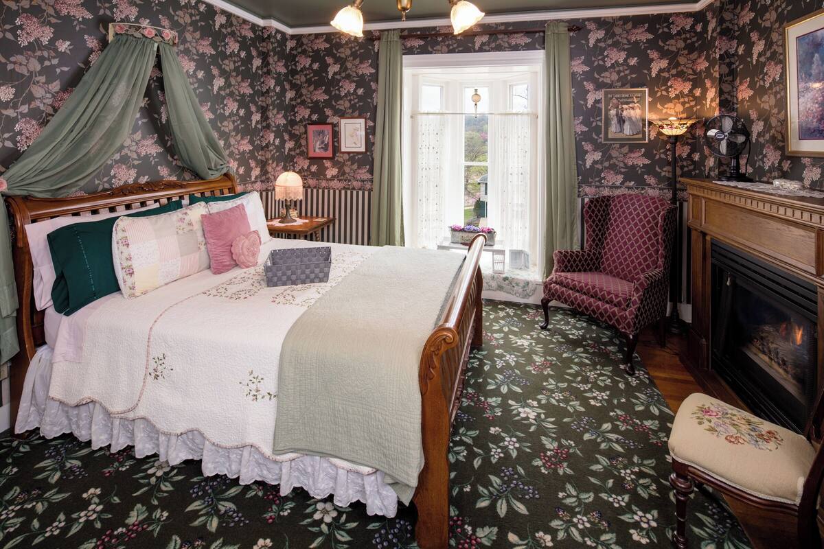 A bedroom with a four-poster bed, a chair, a fireplace, and floral wallpaper.