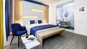 Moderni hotellihuone, jossa on sänky, työpöytä, tuoli, penkki ja lasiseinällä varustettu kylpyhuone.