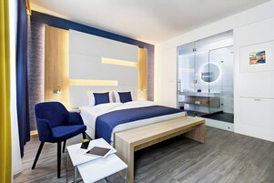 Una habitación de hotel moderna con cama, escritorio, silla, banco y baño con paredes de vidrio.