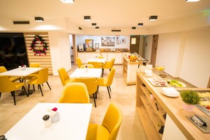 Un comedor moderno con sillas amarillas y mesas blancas, una barra de buffet con comida y bebidas, y un adorno navideño en la pared.