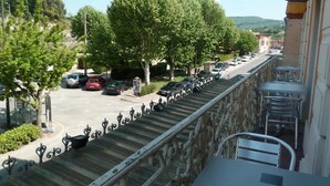 A balcony with a stone balustrade overlooking a street lined with parked cars and trees. - Luberon L'hôtel (Apt)
