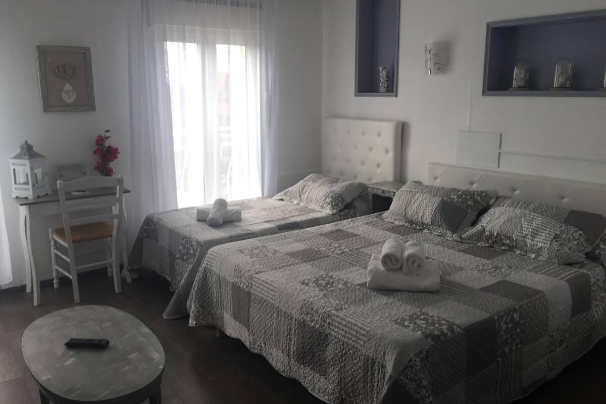 Quarto de hotel com duas camas, uma escrivaninha, uma cadeira e uma mesinha.