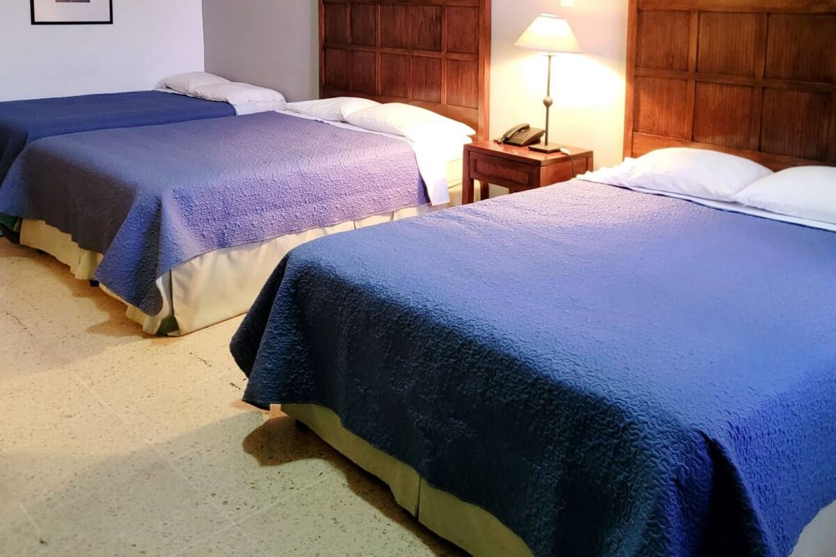 A hotel room with two beds, each with a blue blanket, a wooden headboard, and a bedside table with a lamp.