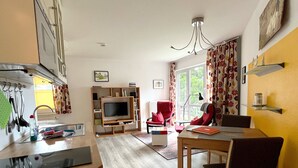 Een moderne keuken met een inbouwoven en magnetron, een eethoek met een houten tafel en stoelen, een rode fauteuil en een televisie in de woonkamer.