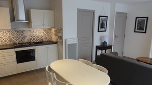 Una cocina compacta con armarios blancos, un horno empotrado y una cocina a gas. En la zona de estar contigua hay una mesa de comedor con sillas y un sofá gris.