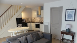 Una sala moderna con un sofá gris, una mesa de centro de madera y una mesa de comedor blanca con sillas. Se ve una cocina con armarios blancos y salpicadero con diseño en el fondo.