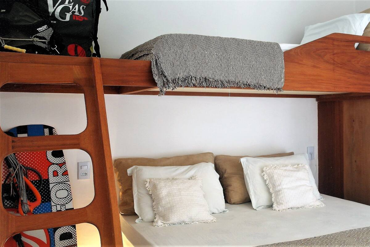 A bunk bed with a wooden frame, a lower bed with white bedding, and a shelf unit with various items.