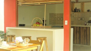Un coin repas avec une table dressée pour deux personnes, un panier en osier rempli de fruits posé sur le comptoir, et une cuisine en arrière-plan.