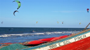 A beach with multiple kites flying, a boat with 'MAR E SOL PREA' written on it, and the ocean in the background. - Pousada SeaWind (Cruz)