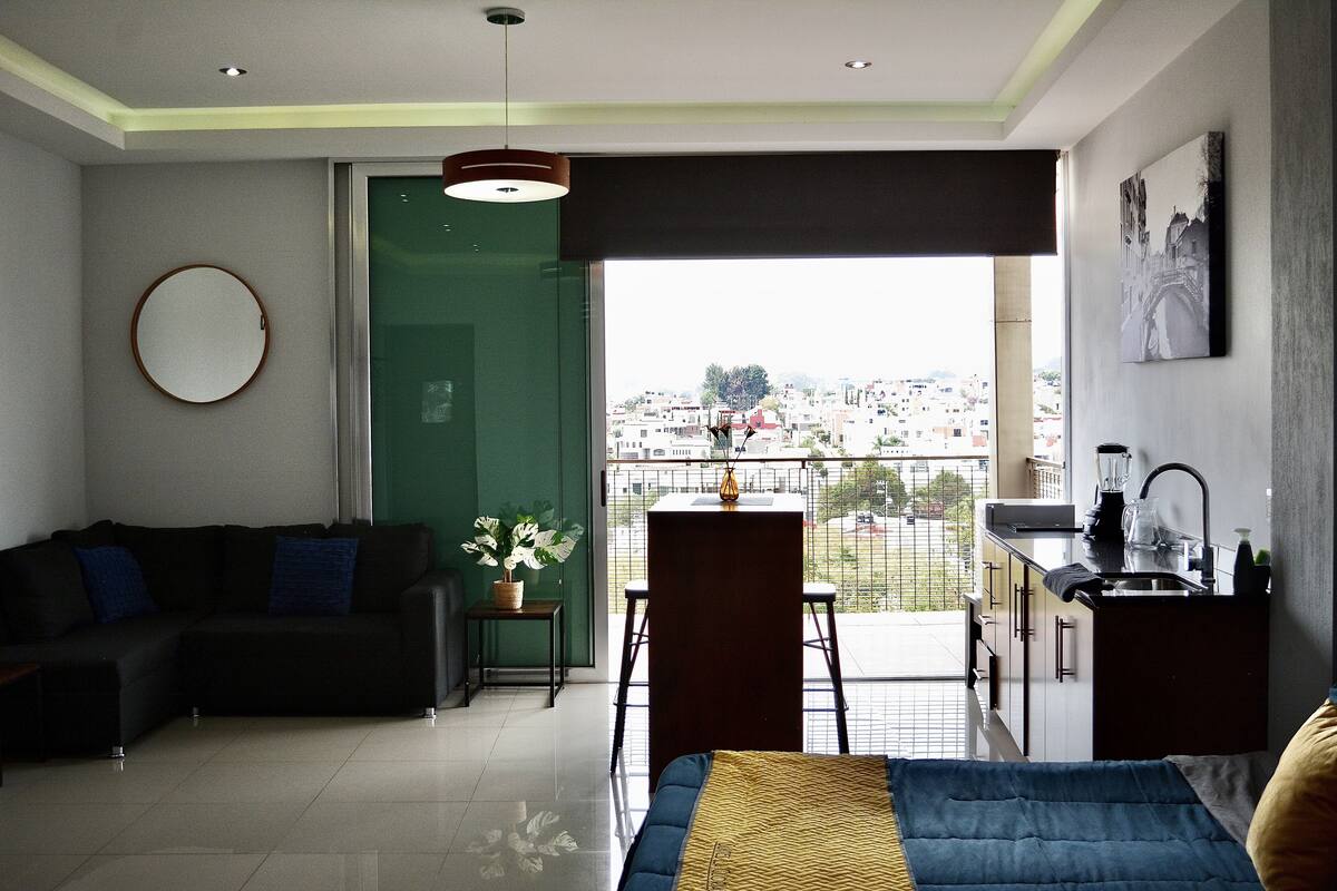 Een moderne woonkamer met een bank, een bed, een kleine keukenhoek en een balkon met uitzicht op de stad.