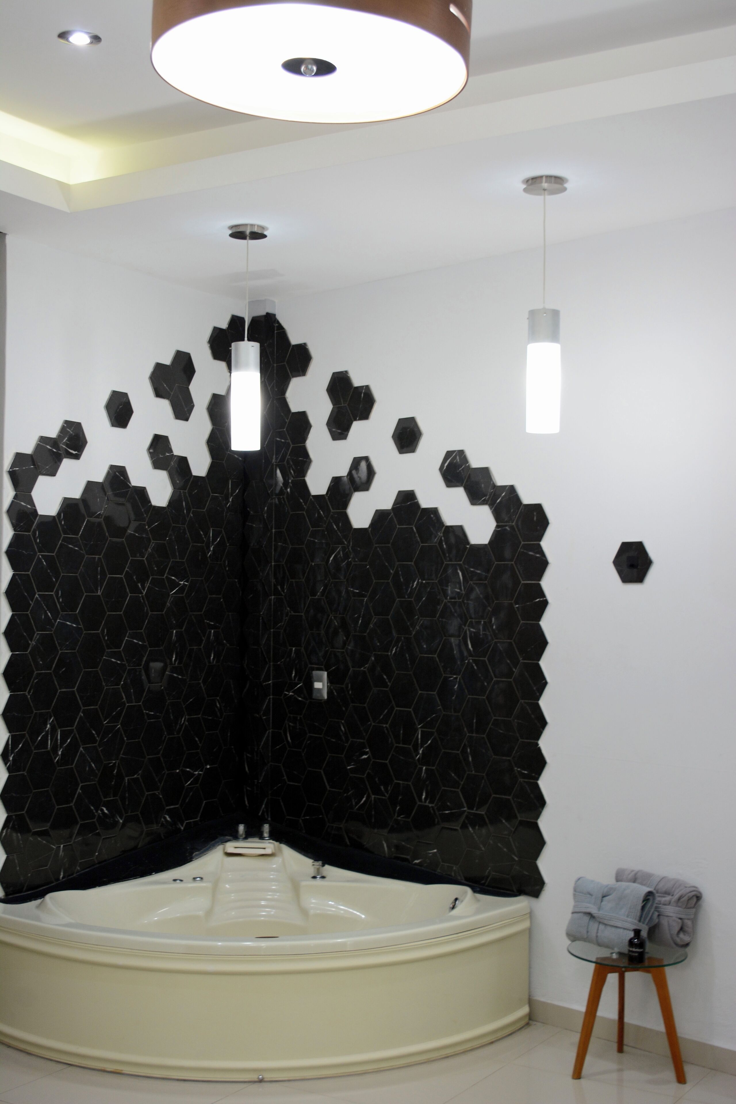 Un baño moderno con pared de azulejos hexagonales negros, una bañera blanca y un taburete de madera pequeño con toallas.