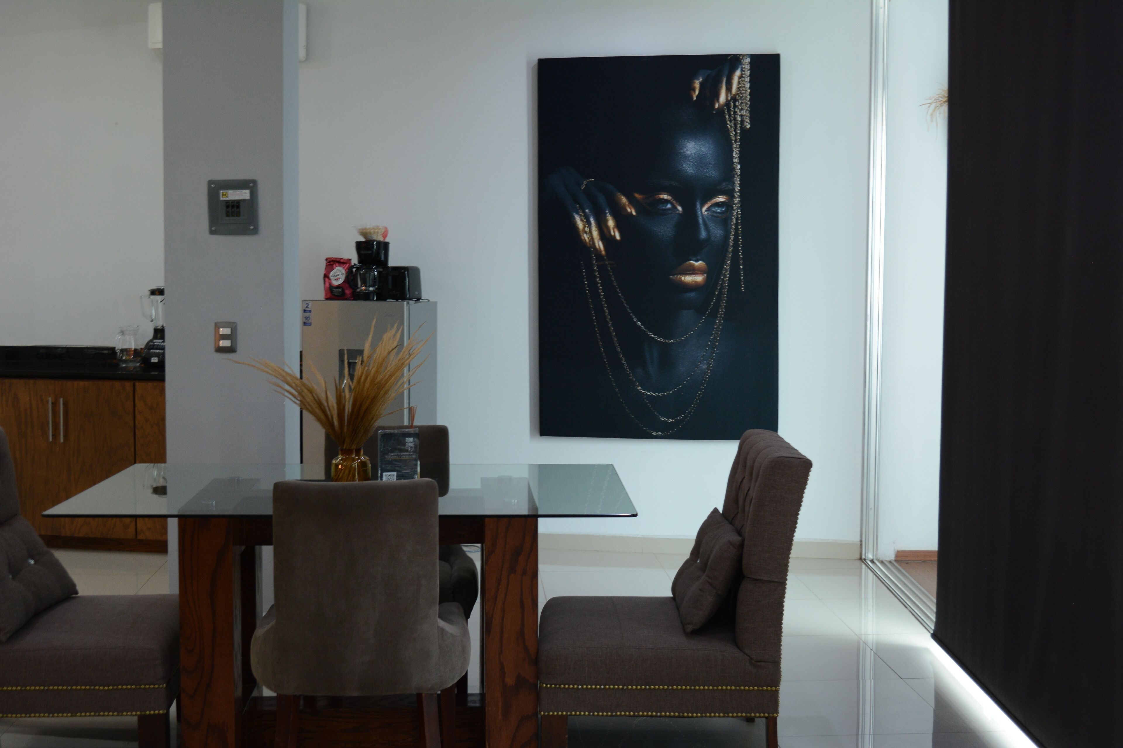 A modern dining area with a glass table, two upholstered chairs, a vase with dried plants, and a large abstract painting on the wall.