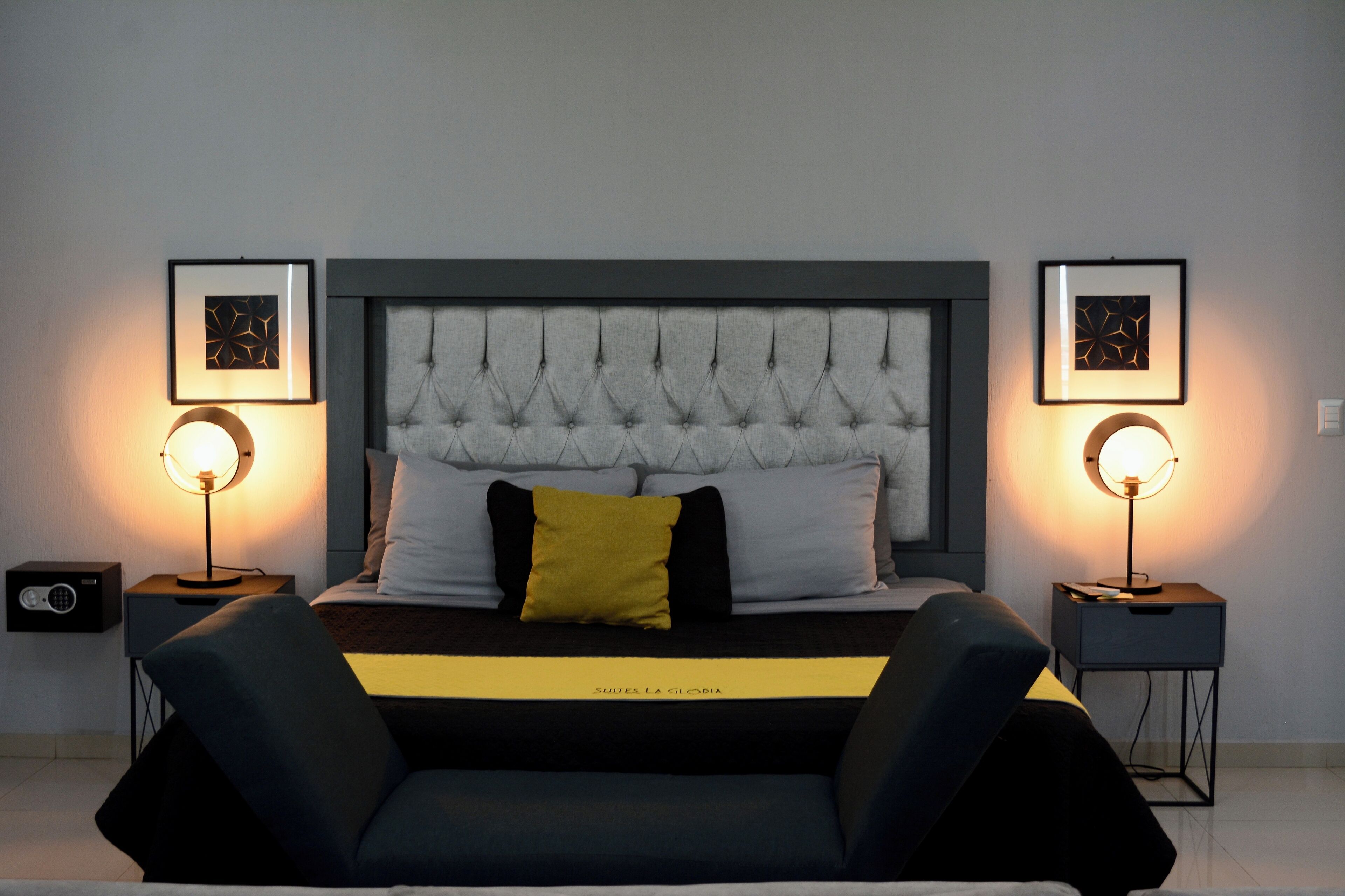 Une chambre à coucher moderne avec une tête de lit capitonnée, un lit recouvert d’une couvre-lit noire et jaune, deux tables de chevet avec des lampes et des œuvres d’art encadrées accrochées aux murs.