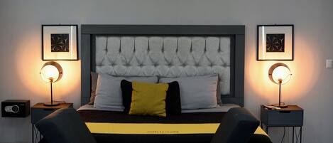 Une chambre à coucher moderne avec une tête de lit capitonnée, un lit recouvert d’une couvre-lit noire et jaune, deux tables de chevet avec des lampes et des œuvres d’art encadrées accrochées aux murs.