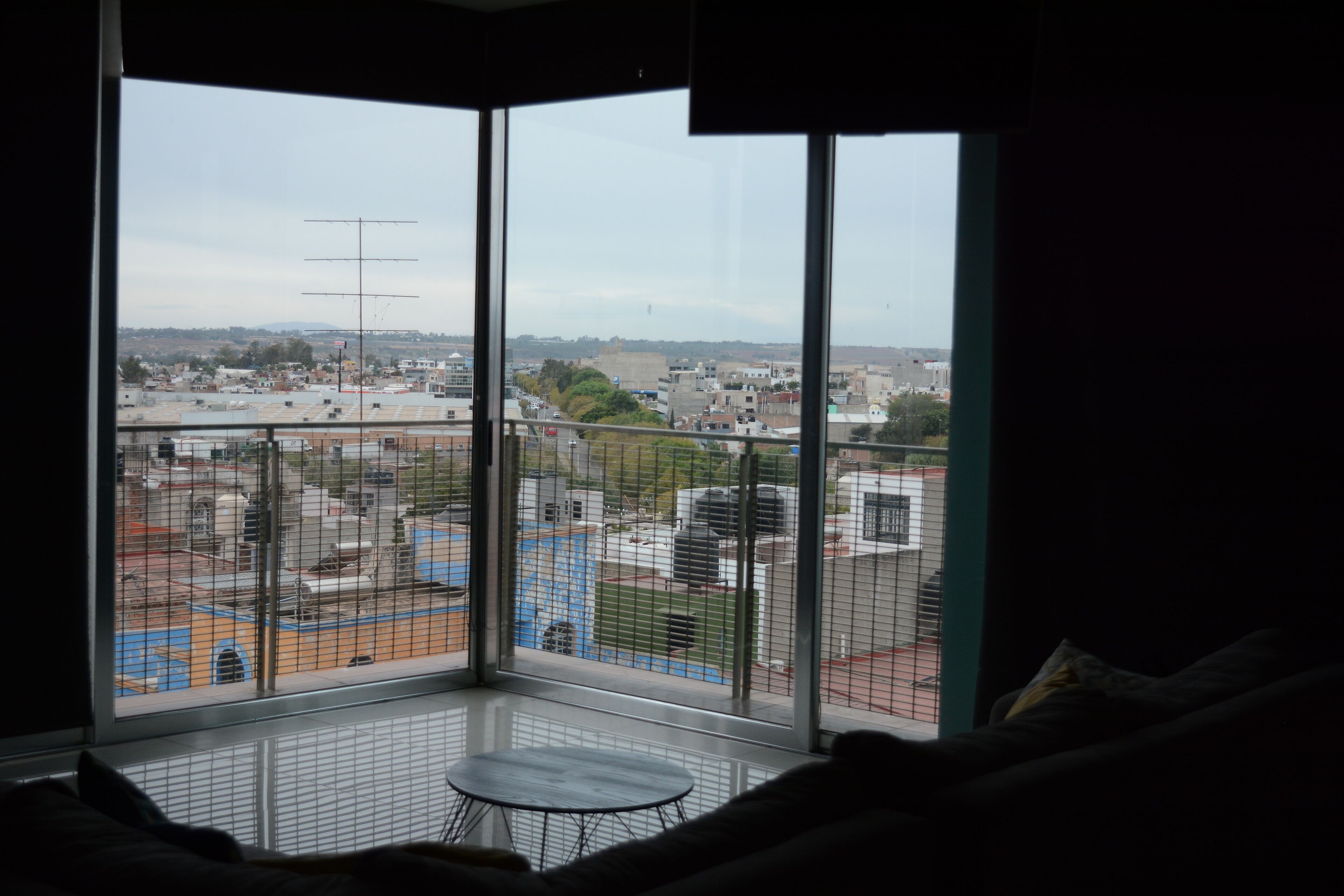 A room with a large window overlooking a cityscape, a small round table, and a bed with a headboard.