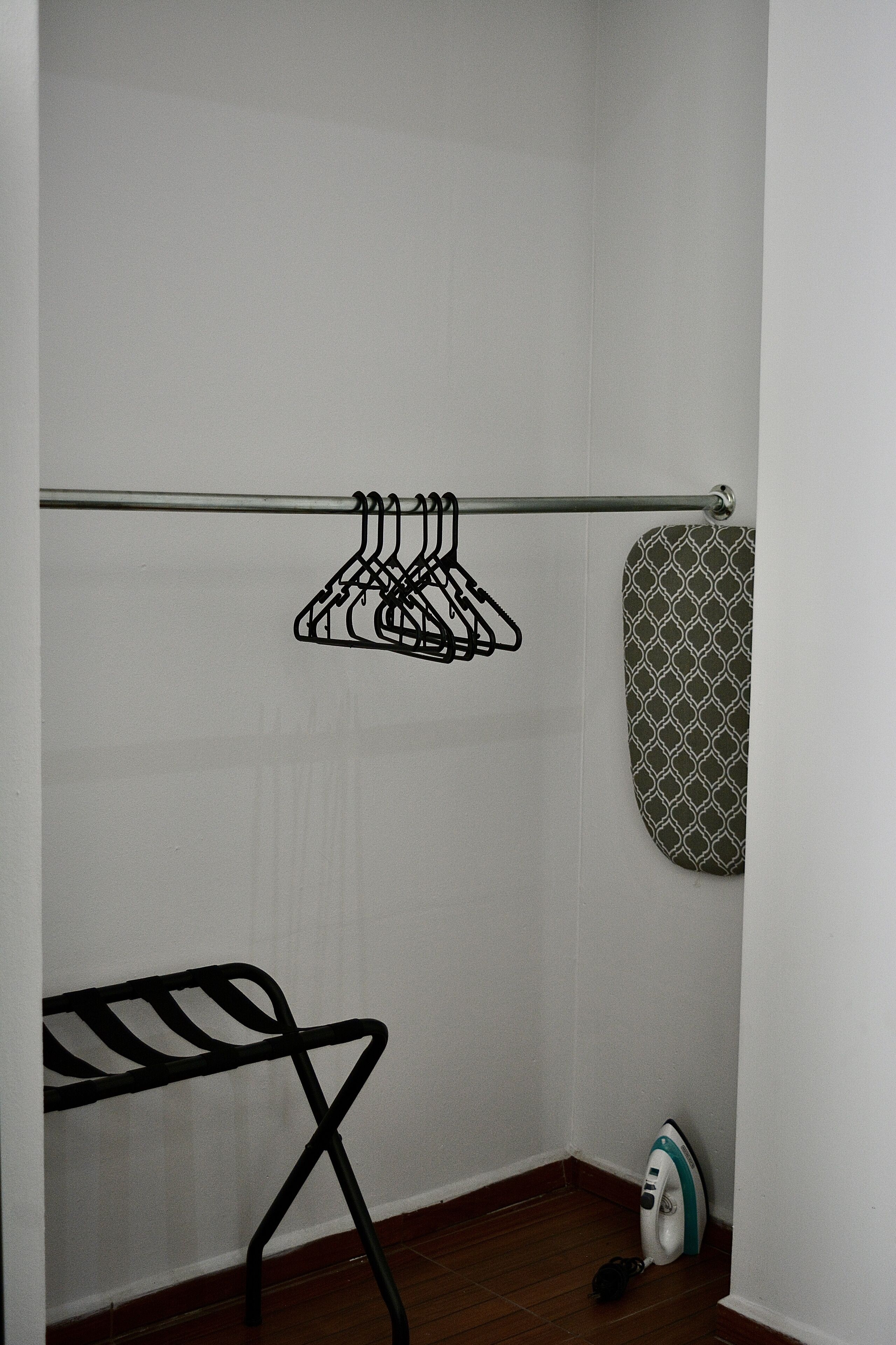 A corner of a room with a metal rack holding clothes, a patterned bath mat, and a black ironing board with an iron.