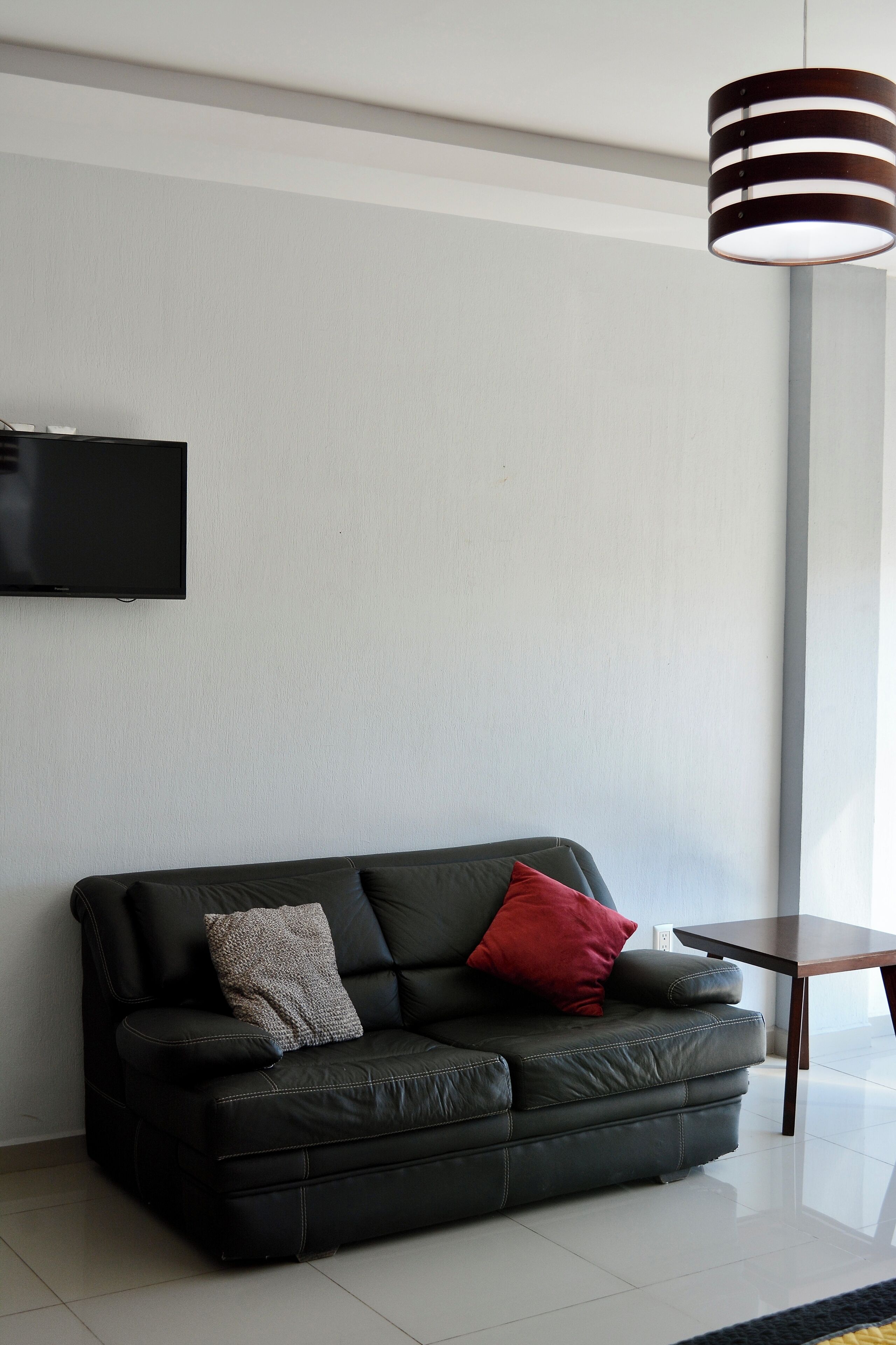 A modern living room with a black leather sofa, a flat-screen TV mounted on the wall, and a hanging pendant light.