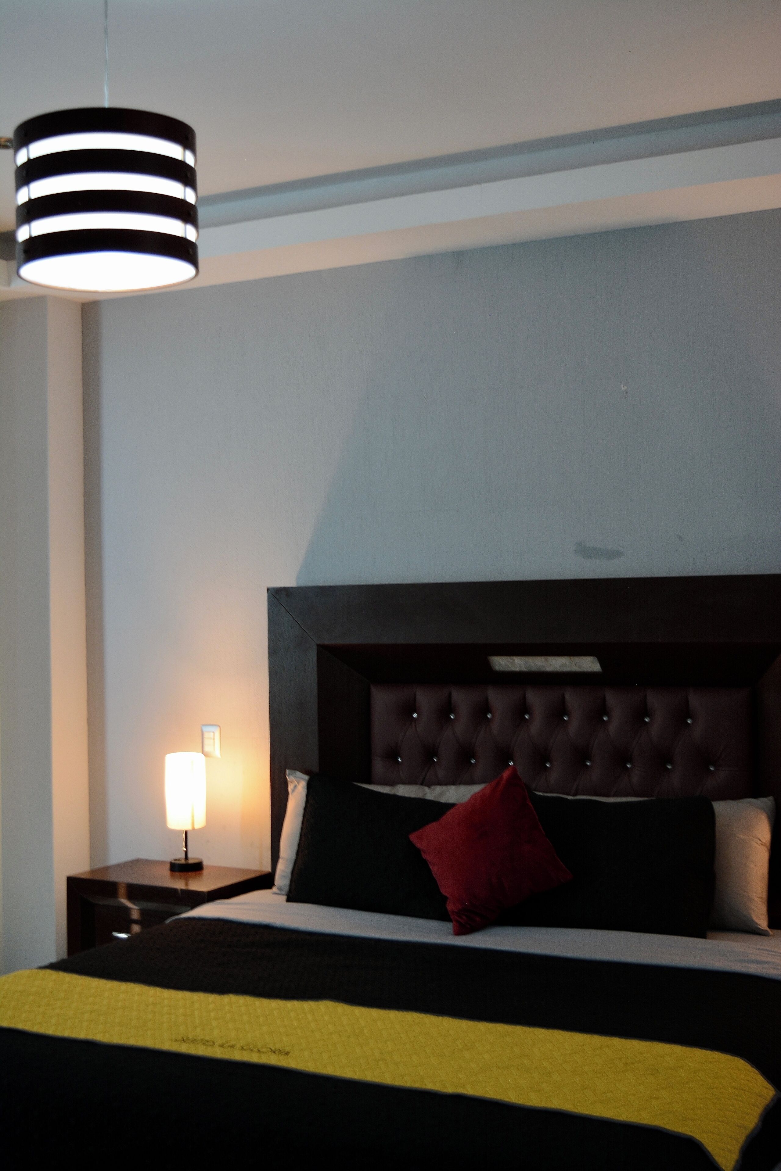 A hotel room with a large bed, a dark headboard, a bedside table with a lamp, and a hanging light fixture.