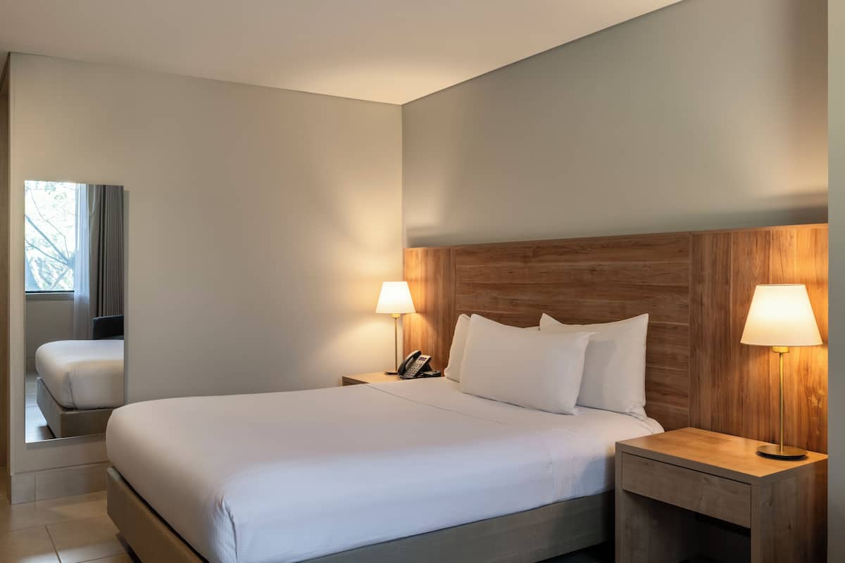 A hotel room with a large bed, wooden headboard, two bedside lamps, a nightstand with a phone, and a window with curtains.