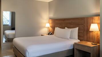 Une chambre d’hôtel avec un grand lit, une tête de lit en bois, deux lampes de chevet, une table de chevet avec un téléphone et une fenêtre avec des rideaux.