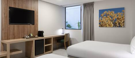 Habitación de hotel con cama, escritorio con silla, televisor montado en un panel de madera, refrigerador pequeño y ventana con cortinas.