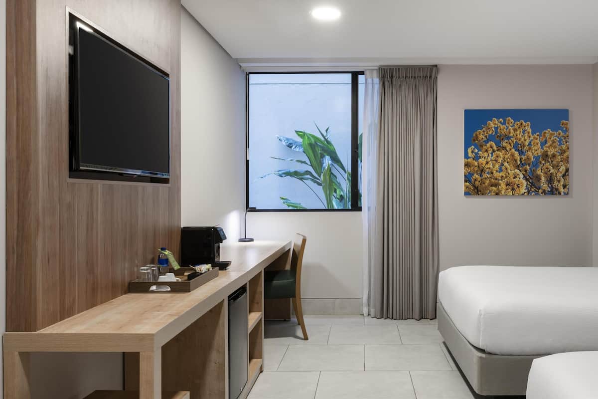 A hotel room with a wooden desk, a flat-screen TV, a bed, a window with a view of plants, and a painting of flowers.