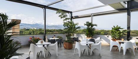 Eine Dachterrasse mit weißen Stühlen und Tischen, Blumentöpfen und Blick auf Berge.