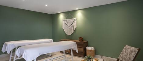 A room with two white beds, a wooden cabinet, a woven chair, and a macrame wall hanging.