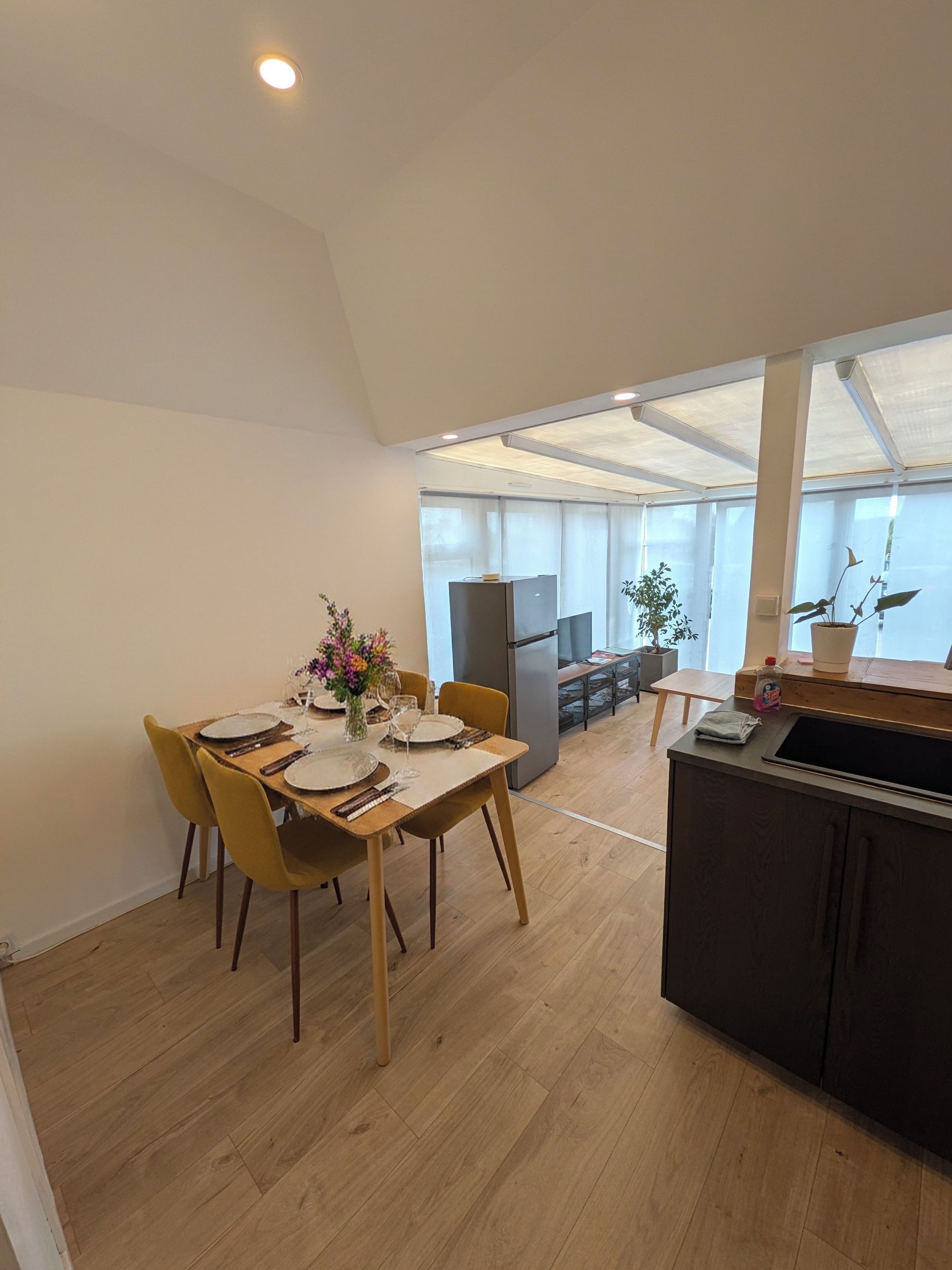 A modern kitchen with a dining area, wooden flooring, and a large window providing natural light.