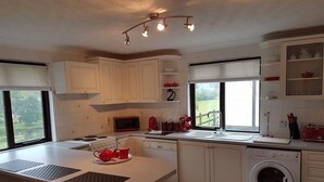 A modern kitchen with white cabinets, a built-in microwave, a red kettle, and a washing machine. There is a window providing natural light and a view of the outdoors. - Lake View Villa - Rosecraddoc Manor (Liskeard)