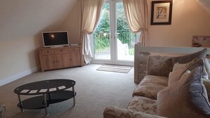 A living room with a sofa, a TV on a wooden cabinet, and a balcony with curtains. - Lake View Villa - Rosecraddoc Manor (Liskeard)