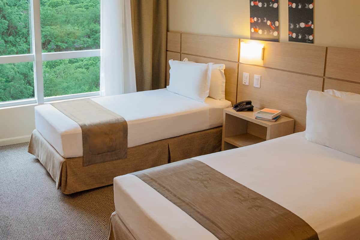 A hotel room with two beds, a nightstand, a lamp, and a view of greenery through the window.
