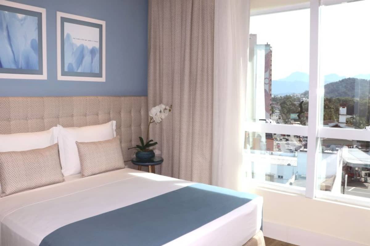 A bedroom with a large bed, a nightstand with a plant, and a window with a city view.