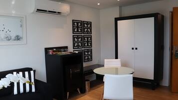 Uma sala de estar moderna com um sofá preto, uma mesa branca e um armário preto. Há um quadro emoldurado na parede e um aparelho de ar condicionado acima do sofá.