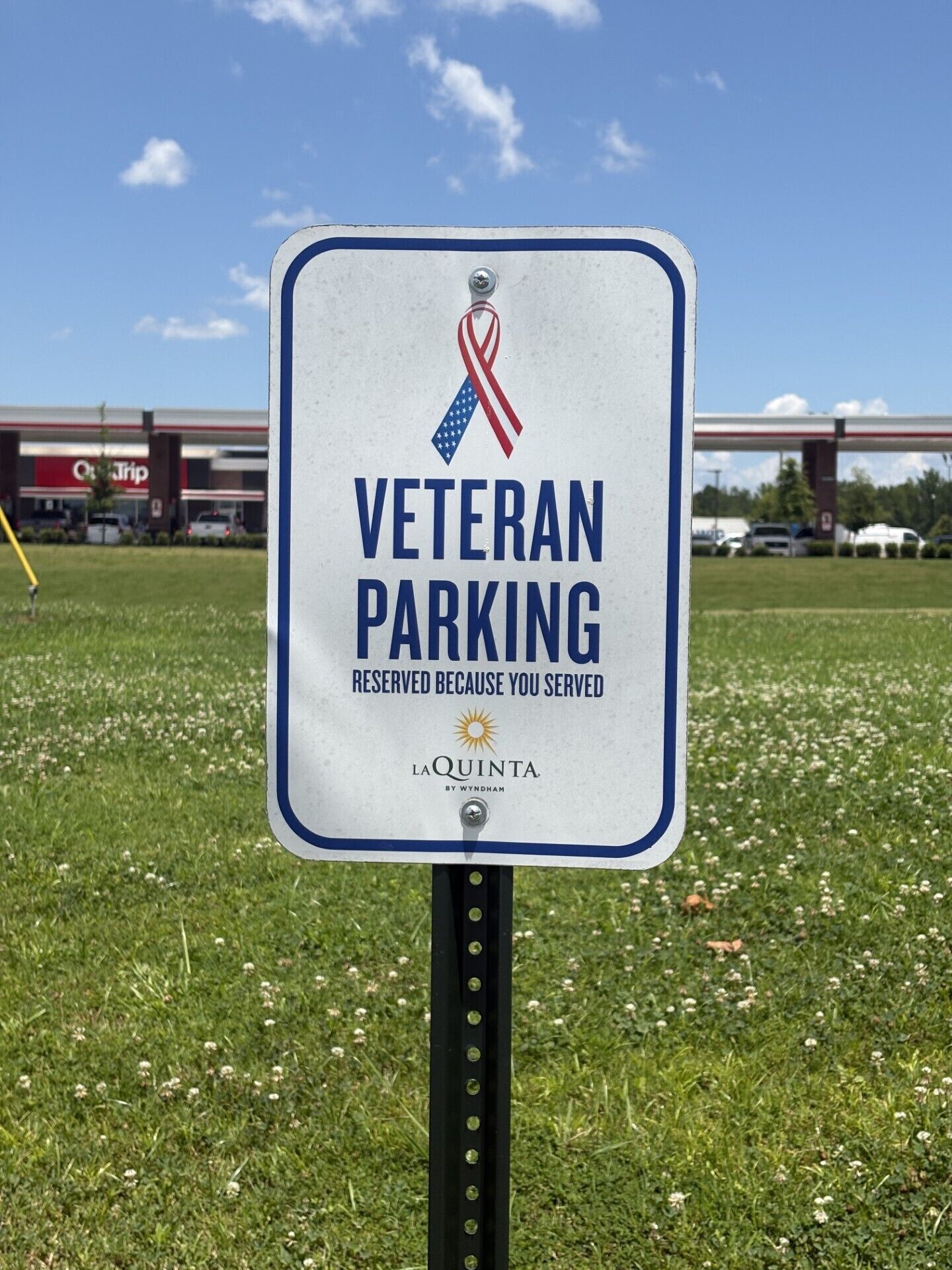 Schild für Parkplätze für Veteranen mit einem Band-Symbol, in einer grünen Umgebung gelegen, mit einer Tankstelle im Hintergrund.