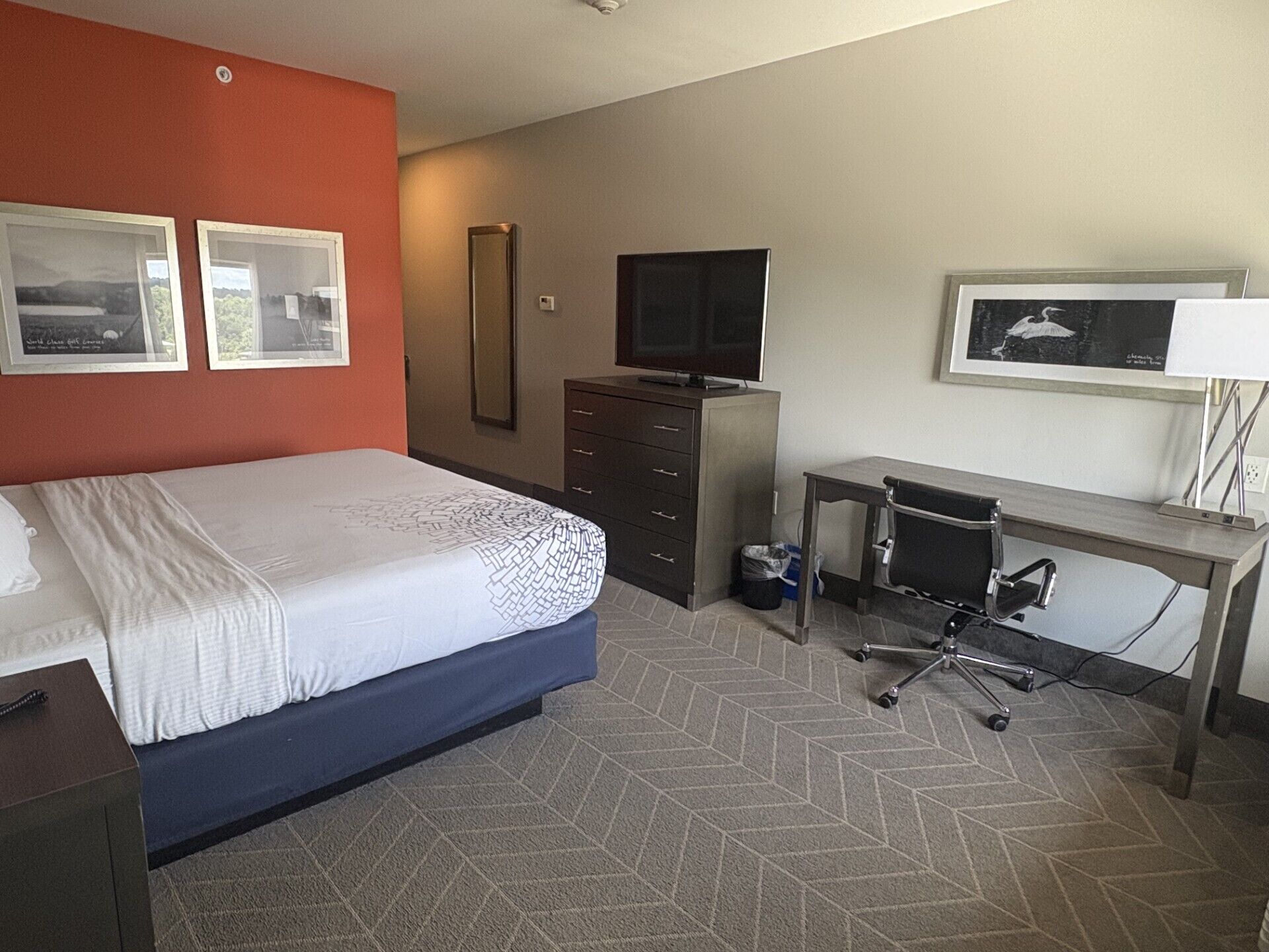 A hotel room with a bed, a desk, a chair, a TV, and two framed pictures on the wall.
