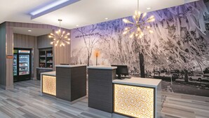 A modern reception area with a mural of trees and a quote on the wall, a reception desk, and a refrigerated display case. - La Quinta Inn & Suites by Wyndham Opelika Auburn (Opelika)