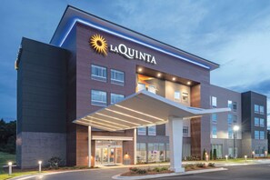 Een La Quinta hotel met een modern design, herkenbaar aan een opvallend zonlogo, een overdekte ingang en een goed verlichte gevel.